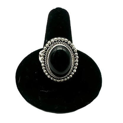 A silver-toned ring with an oval black cabochon stone, displayed on a black velvet ring stand against a white background.